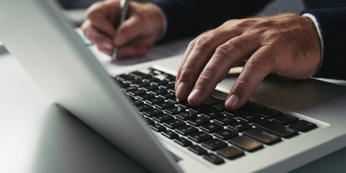 Mãos de uma pessoa utilizando o teclado de um notebook enquanto faz anotações