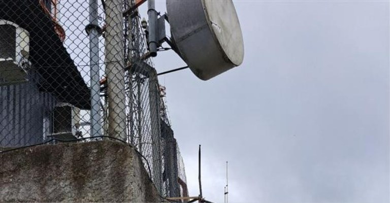 Detalhe de rádio interrompida pela equipe de fiscalização da Anatel. A imagem mostra uma estrutura cercada por uma grade de arame. Há uma grande antena posicionada na parte superior.