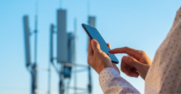 Em primeiro plano, a mão de uma pessoa toca na tela de um celular. Ao fundo, antenas no topo de uma torre de telecom.