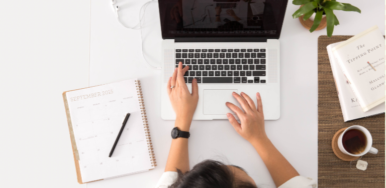 De uma visão vertical, temos uma pessoa a acessar o laptop. Na mesa, há uma agenda com caneta do lado esquerdo e à direita um copo de café e livros. Um pouco à frente do laptop, uma planta de vaso na mesa.