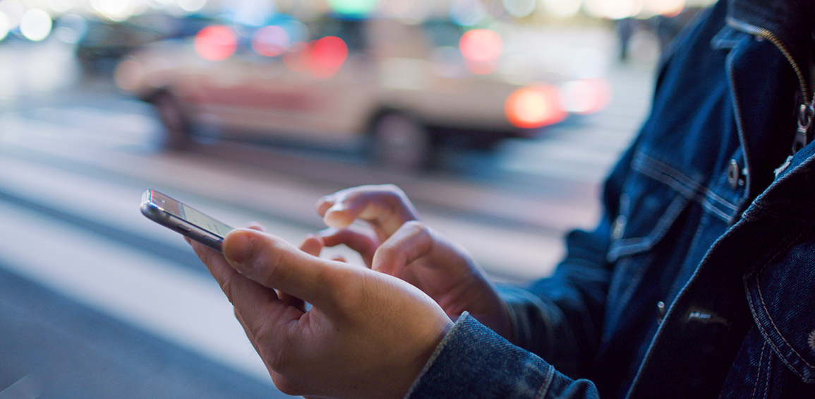 Homem na calçada digitando no celular com faixa de pedestres e carros ao fundo.