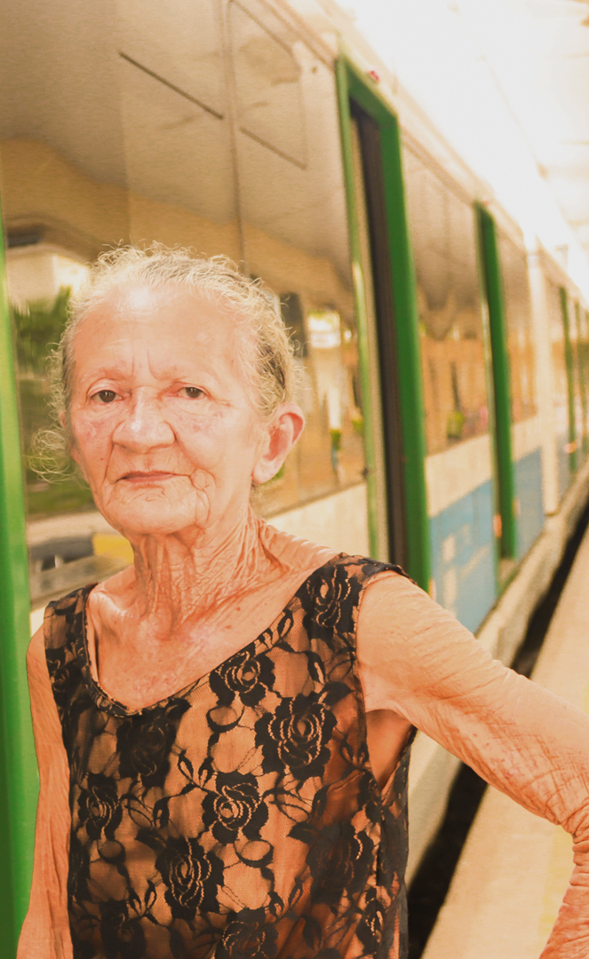 Foto de uma Mulher idosa numa estação de transporte urbano ao lado de um Veículo Leve sobre Trilhos (VLT).