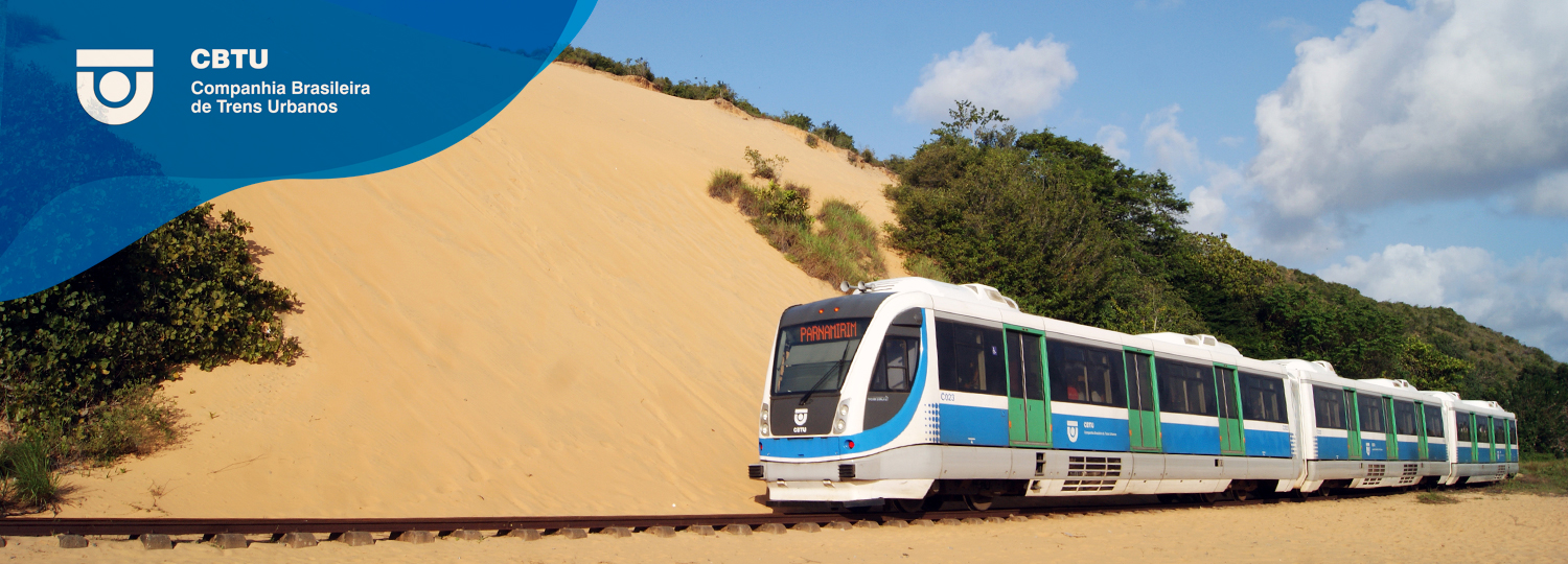 Trem passando sob trilhos. Duna de areia, vegetação, céu azul com nuvens.