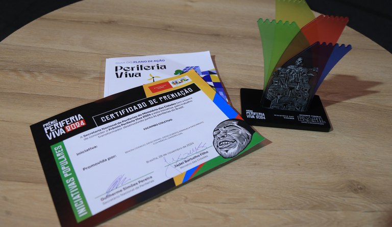 Na foto, sobre uma mesa, o certificado de premiação do Prêmio acima do guia do Plano de Ação e o troféu Periferia Viva ao lado