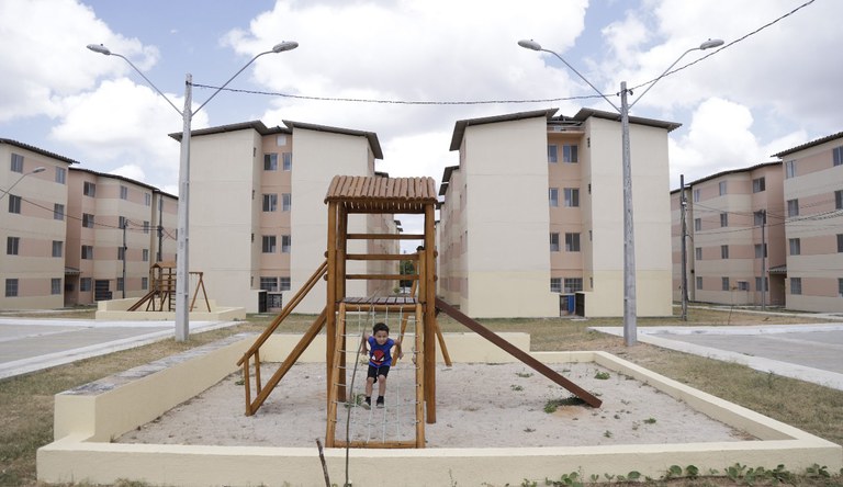 Na foto menino brincando em um brinquedo de rede de cordas no parquinho de um residencial do MCMV