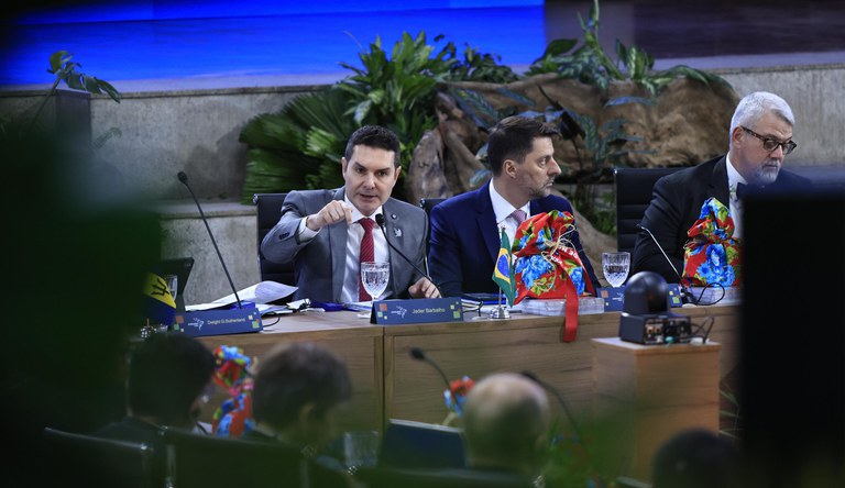 Na foto o ministro das cidades fala sentado a mesa junto a outros