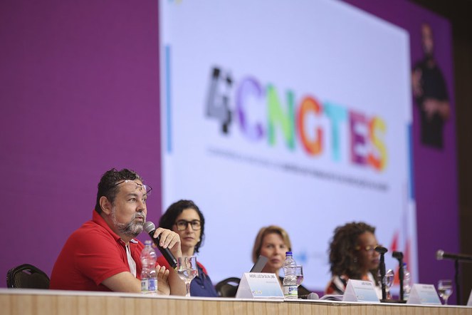 Foto: Thiago Lustosa Fotografia. - quatro participantes do eixo 1 da 4ª CNGTES sentados à mesa no palco. Atrás deles, a logomarca da 4ª CNGTES