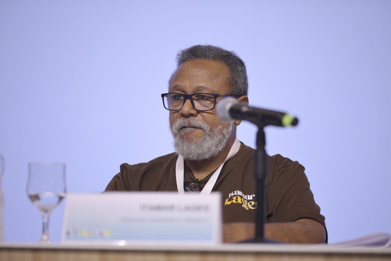 Foto: Thiago Lustosa Fotografia -  Itamar Lages, um homem negro, de cabelos e barba grisálias, ele esta sentado e fala em frente ao microfone suspenso à mesa. Ele usa óculos e veste uma blusa marrom.