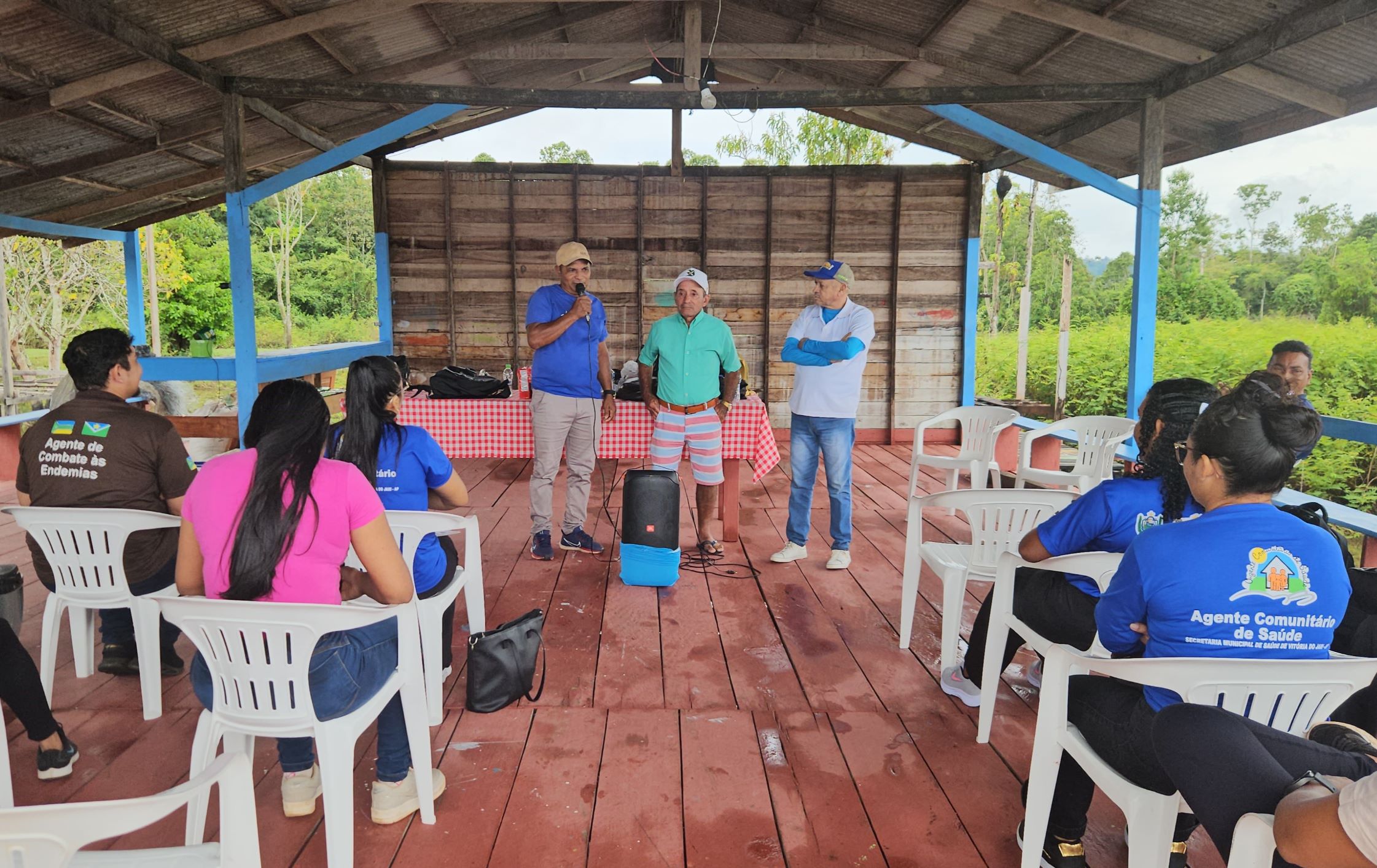 Grupo em reunião sob telhado de madeira, com três homens em pé conversando com pessoas sentadas.