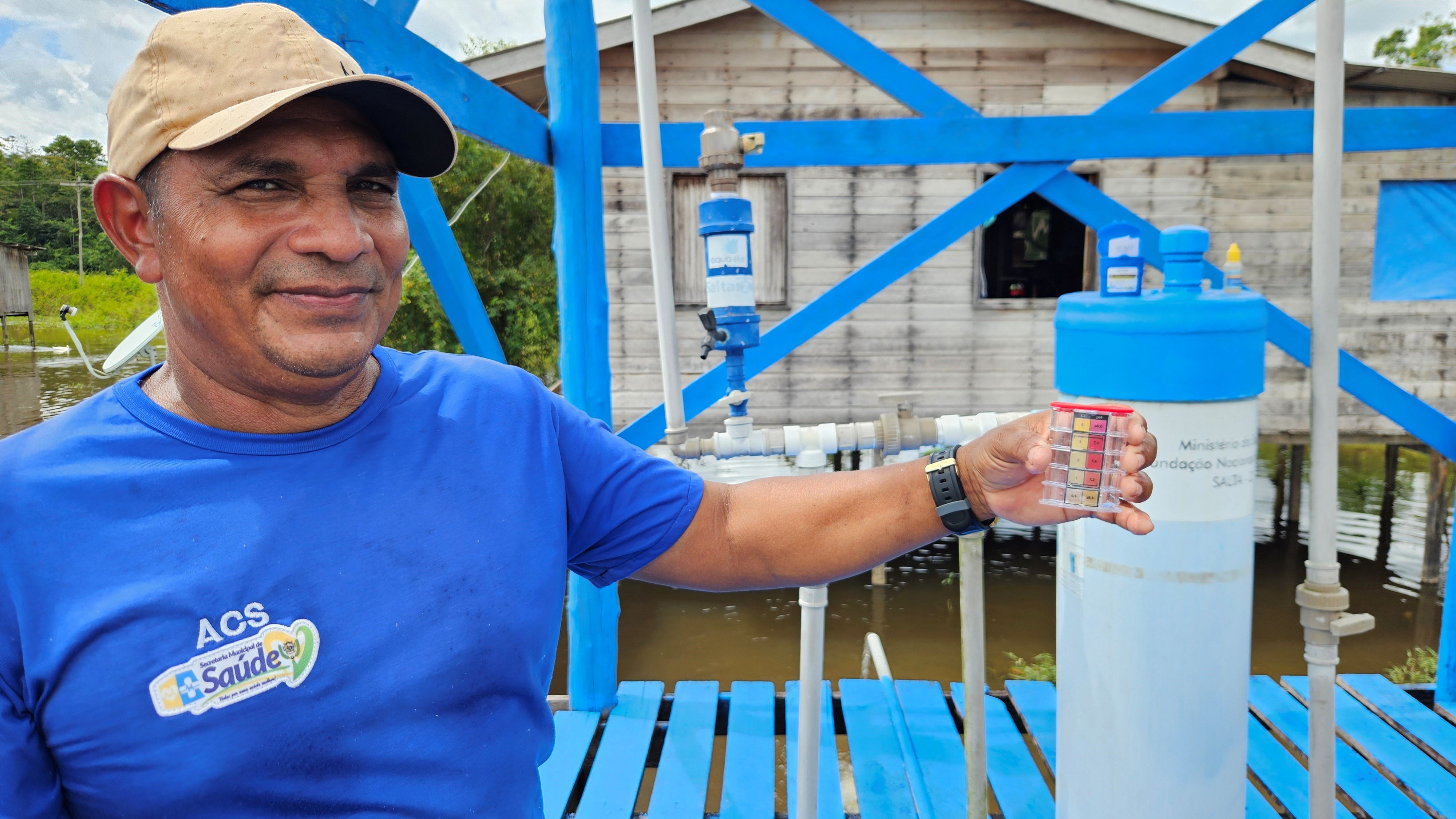 Homem de boné e camiseta azul sorri diante de equipamento de água, segurando um medidor químico.