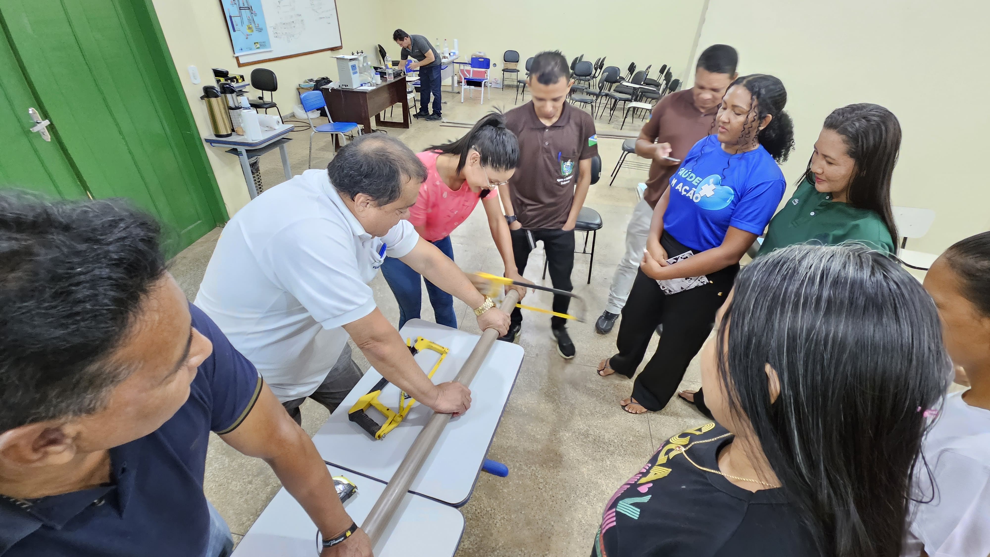 Pessoas em oficina acompanham o corte manual de um cano de PVC marrom feito com arco de serra.