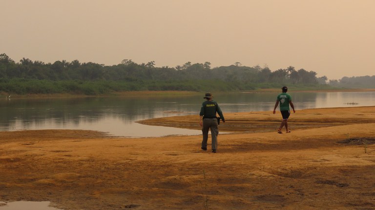 A imagem mostra duas pessoas caminhando em uma área de areia à margem de um rio Tapajós ou Xingu, cercado por vegetação densa. Uma delas veste um colete com "Ibama" nas costas, enquanto a outra usa uma camisa com o texto "monitor". O cenário parece calmo, ao entardecer.