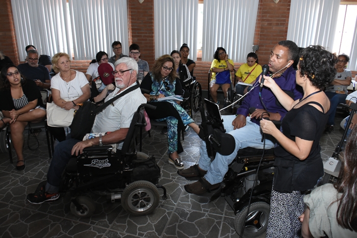 Secretário Marco Pellegrini da SNPD em cadeira de rodas motorizada palestra ao microfone. Na sala, pessoas com e sem deficiência. Ele é negro, cabelos curtos e grisalhos. Veste calça jeans, camisa azul e sapatos.   Foto: Luciana Gifone - SEDPcD