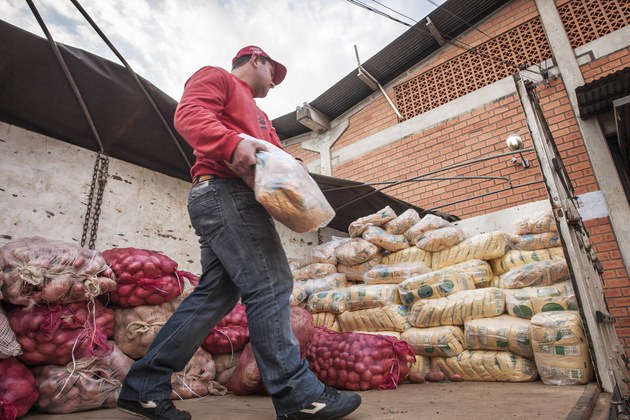 Homem de calça jeans, sapato preto, blusa vermelha e boné vermelho, carregando cestas de alimentos básicos.