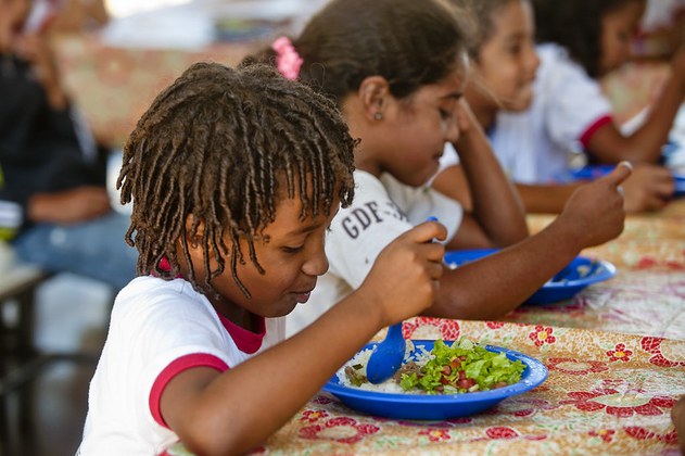 A foto representa crianças se alimentando, elas usam uniformes escolar branco com vermelho, o prato é azul onde contem a comida é azul e contem arroz, carne, feijão e salada.