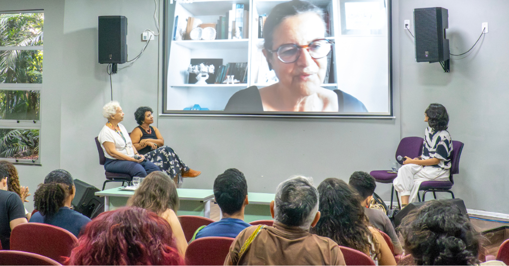 quatro mulheres, uma em vídeo conferencia, conversam em palco de auditório