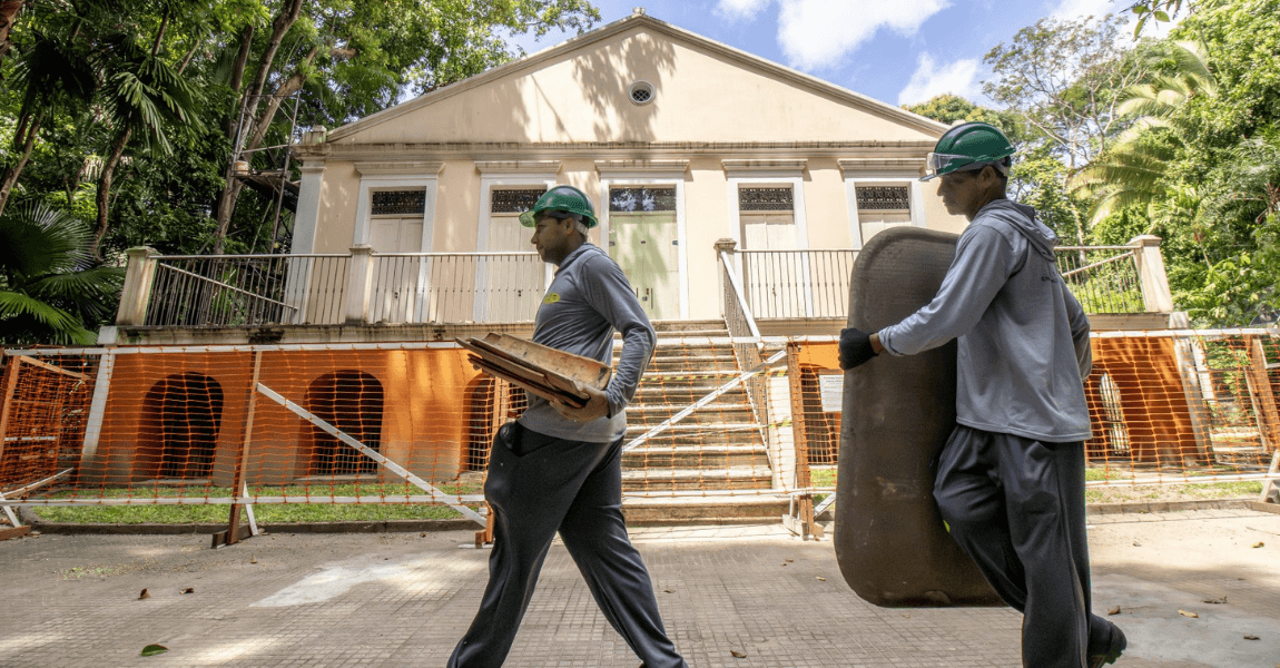 Dois pedreiros com calças, camisa de manga longa e capacete carregam material de construção civil diante do prédio histórico chamado de Rocinha
