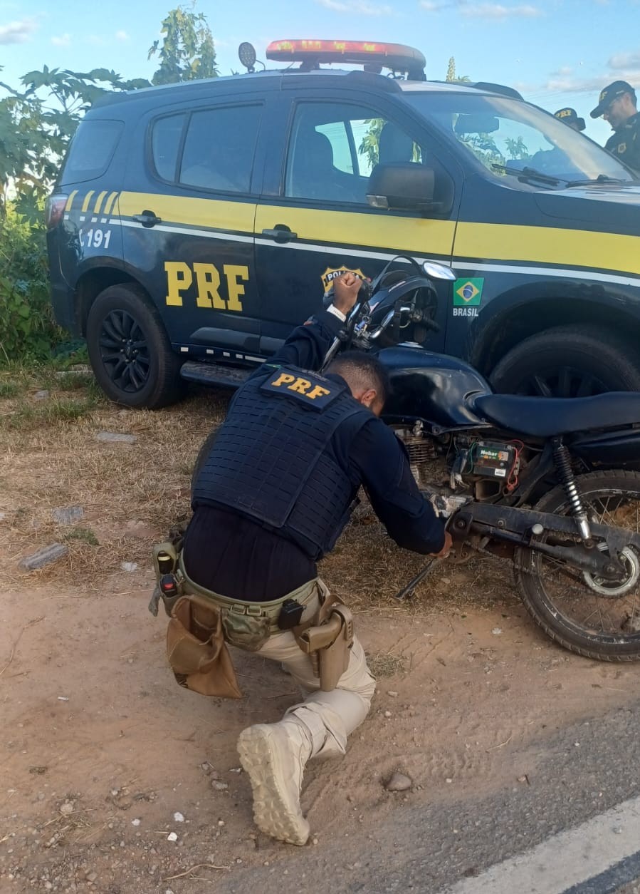 Um PRF examinando a motocicleta adulterada. Ao fundo uma viatura.