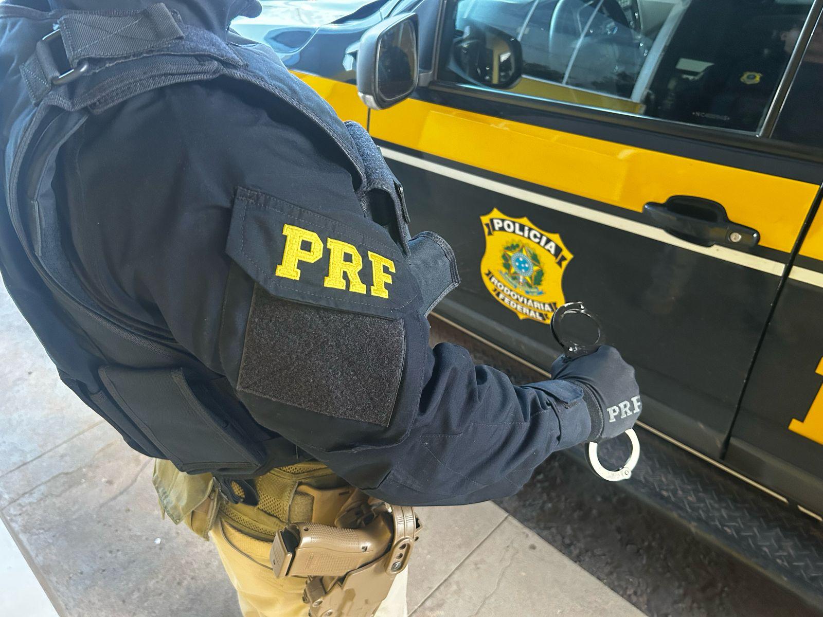 Imagem em ambiente externo mostrando um policial rodoviário federal realizando a conferência de um documento veicular. Em primeiro plano, o agente, trajando uniforme operacional da PRF, segura com as duas mãos uma folha impressa contendo informações e códigos de identificação típicos de documentos oficiais de trânsito. Ao fundo, observa-se o capô de uma viatura da Polícia Rodoviária Federal, com destaque para o brasão institucional e a sigla “PRF” em letras amarelas sobre a pintura preta característica da corporação. O enquadramento enfatiza a atividade de fiscalização documental, representando procedimentos de verificação e controle realizados pela PRF para garantir a regularidade de veículos e condutores nas rodovias federais.