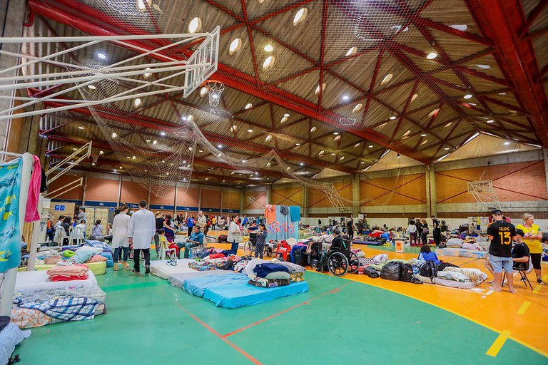 A imagem retrata um ginásio esportivo interno que foi convertido em uma área temporária de abrigo e acomodação em Porto Alegre. Várias camas e roupas de cama estão dispostas no chão, criando áreas para dormir. O ginásio é espaçoso, com um teto alto com vigas de metal expostas e luminárias. Redes pendem do teto, possivelmente para atividades esportivas como vôlei ou basquete, indicando a função original do local antes da conversão. Pessoas estão espalhadas pelo espaço; algumas estão em pé, conversando ou se movimentando, enquanto outras estão organizando suas áreas nas camas. .