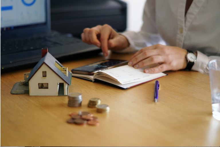 Miniatura de uma casa com algumas moedas e uma mulher fazendo cálculos
