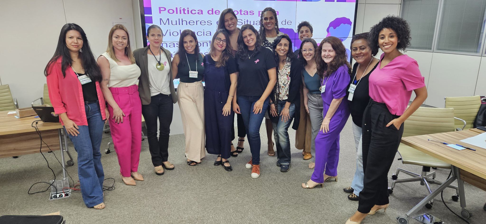 Em uma sala, um grupo de mulheres participantes da oficina em pé e sorrindo para a foto. Ao fundo uma projeção de slides escrito "Política de cotas para Mulheres".