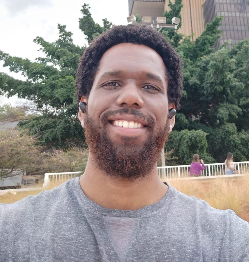 Homem negro sorrindo, com barba, bigode e cabelos pretos crespos. Utiliza fones de ouvido sem fio. Veste uma camiseta cinza. No fundo, um prédio e uma árvore.