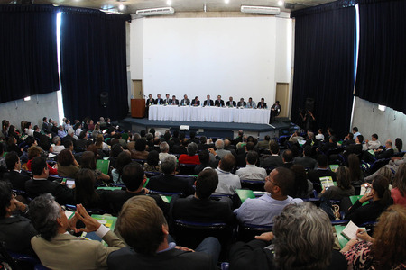 Imagem de uma sala lotada de pessoas. Todas estão sentadas e estão olhando para as pessoas que estão sentadas na bancada da frente.