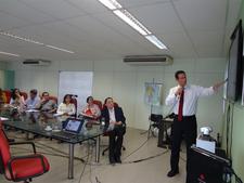 Várias pessoas numa sala, sentadas e assistindo a outra que está na frente discursando.