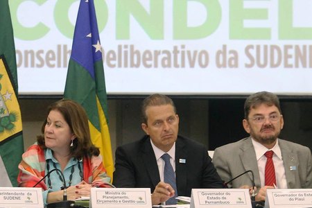 Imagem de Eduardo Campos sentado atrás de uma bancada com mais duas pessoas. Atrás deles, um painel escrito: CONDEL, Conselho Deliberativo da Sudene.