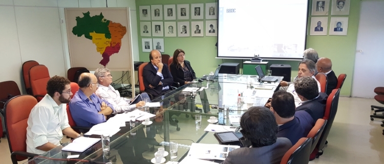 A foto retrata uma reunião com várias pessoas em torno de uma mesa de vidro, falando sobre uma projeção.