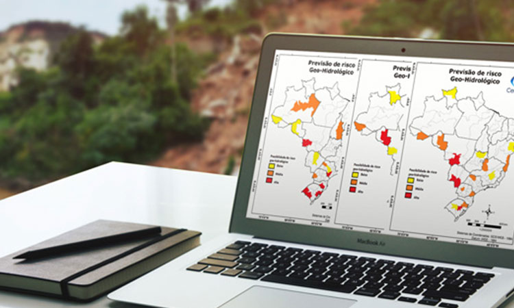 Computador em cima de uma mesa com imagens de mapas/monitoramento. Por trás do computador, tem a imagem de um morro, com risco de deslizamento.