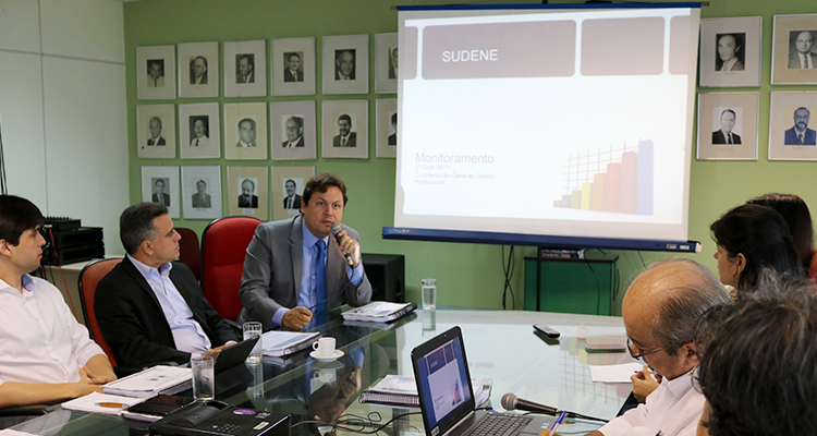 A foto traz o superintendente Marcelo Neves falando durante o ciclo de monitoramento de projetos da Sudene. Há uma projeção de gráficos em um painel e pessoas anotando dados. Todos estão numa sala. Há seis pessoas na foto.