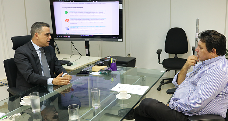 A foto retrata duas pessoas conversando: o diretor de Planejamento da Sudene, Alexandre Gusmão, e o prefeito de João Câmara (RN), Maurício Caetano Damacena. Ambos estão sentados próximos a uma mesa e acompanham apresentação em um monitor de TV.