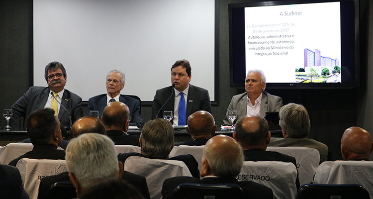 Superintendente Marcelo Neves discursa em mesa de abertura de evento sobre indústrias no Nordeste. Está acompanhando por outros três palestrantes e se dirige a uma plateia. Há pessoas sentadas em um auditório e acompanham apresentação. 