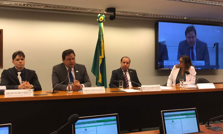 Superintendente da Sudene sentado à mesa com mais três pessoas, durante pronunciamento na Comissão de Integração Nacional, Desenvolvimento Regional e da Amazônia.Ao fundo, a bandeira do Brasil e um telão com a imagem de Marcelo Neves.