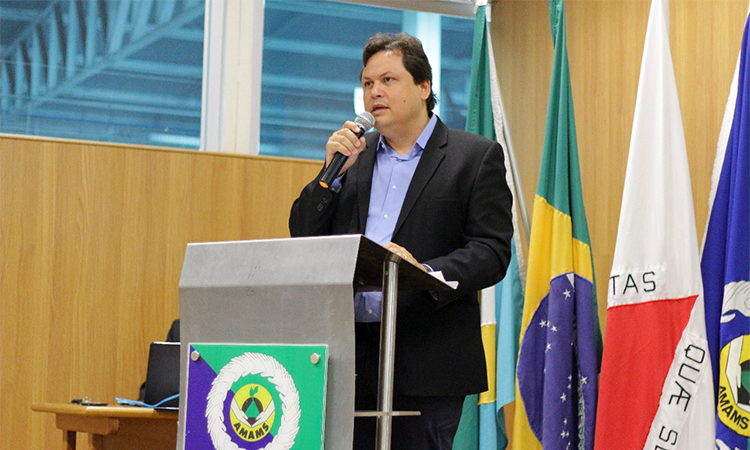 Foto do superintendente Marcelo Neves realizando apresentação a uma plateia. Ele está em um palco, em pé atrás de um púlpito e aponta para um telão. Há pessoas em um andar inferior assistindo a palestra.