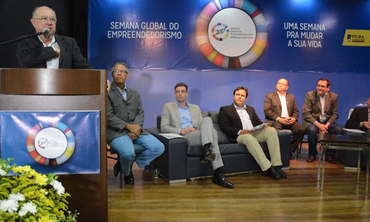 A foto mostra o superintendente Marcelo Neves no palco do evento, sentado ao lado de outras cinco pessoas. Um pouco a frente, em um púlpito, está o prefeito de Feira de Santana, durante seu discurso.