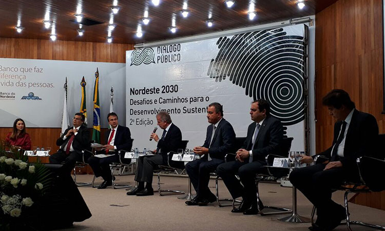 A foto mostra as autoridas no palco, sentadas lado em lado em cadeiras. Não tem mesa. Atrás das autoridades aparecem quatro bandeiras e um painel com o nome do evento.