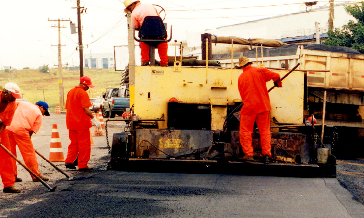 Foto de uma equipe de obras asfaltando uma rua. Há uma máquina com um rolo compressor nivelando a estrutura da pista e trabalhadores ao redor da obra.