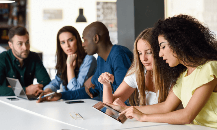 A foto mostra um grupo de estudantes durante um estudo coletivo. São cinco pessoas que estão em uma mesa com materiais escolares e tablets. Eles conversam entre si.