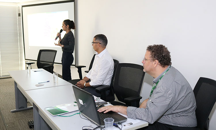 Foto de uma palestrante próxima ao telão e da mesa de duas pessoas sentadas à mesa que fica no palco