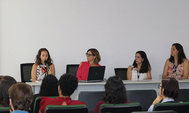 Foto das quatro palestrantes sentadas à mesa e de parte da plateia.