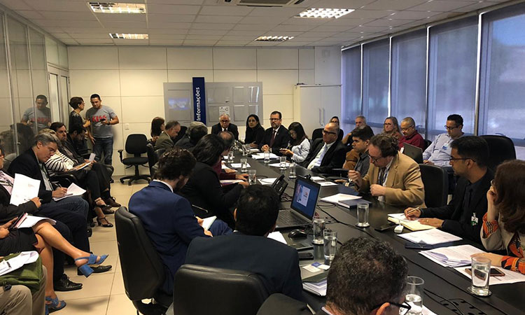 Foto da sala de reunião, com diversas pessoas sentadas à mesa e em cadeiras proximas à mesaatéria