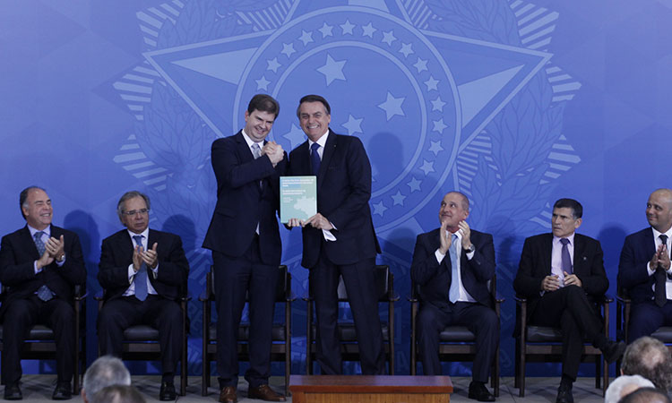 Jair Bolsonaro e Gustavo Canuto em pé, no palco, segurando o documento referente à PNDR. Na parte de trás aparecerm outras lideranças sentadas. 