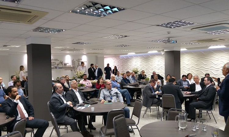 A foto mostra os participantes da reunião sentados às mesas. São diversas mesas redondas, em um local amplo e fechado.