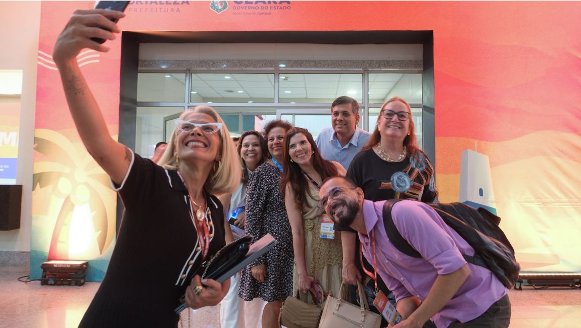 Grupo de sete pessoas sorri para uma selfie na entrada de uma feira de turismo. À esquerda, uma mulher segura o celular com o braço estendido. Alguns participantes usam crachás, e o painel ao fundo exibe as marcas da Prefeitura de Fortaleza e do Governo do Ceará.