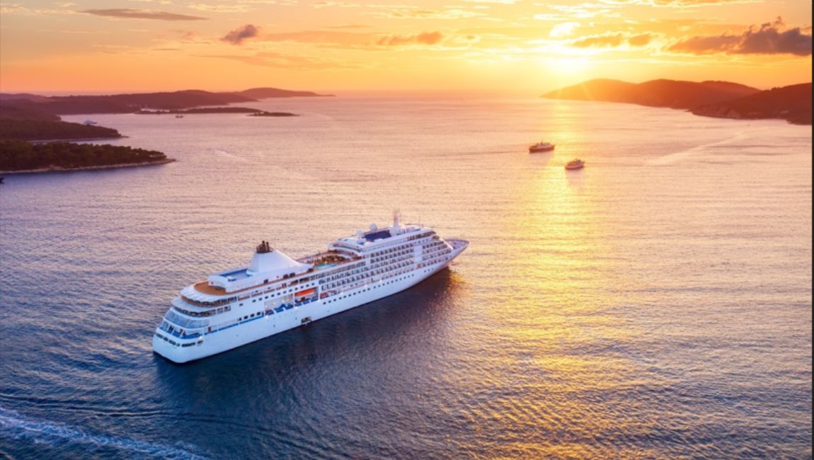 Um navio de cruzeiro navega pelo mar ao pôr do sol. O céu está alaranjado e dourado, com ilhas ao fundo e outros barcos ao longe.